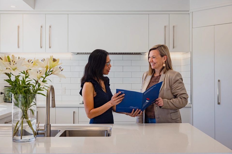 Two women standing at a kitchen bench reading a blue Pukeko management folder.