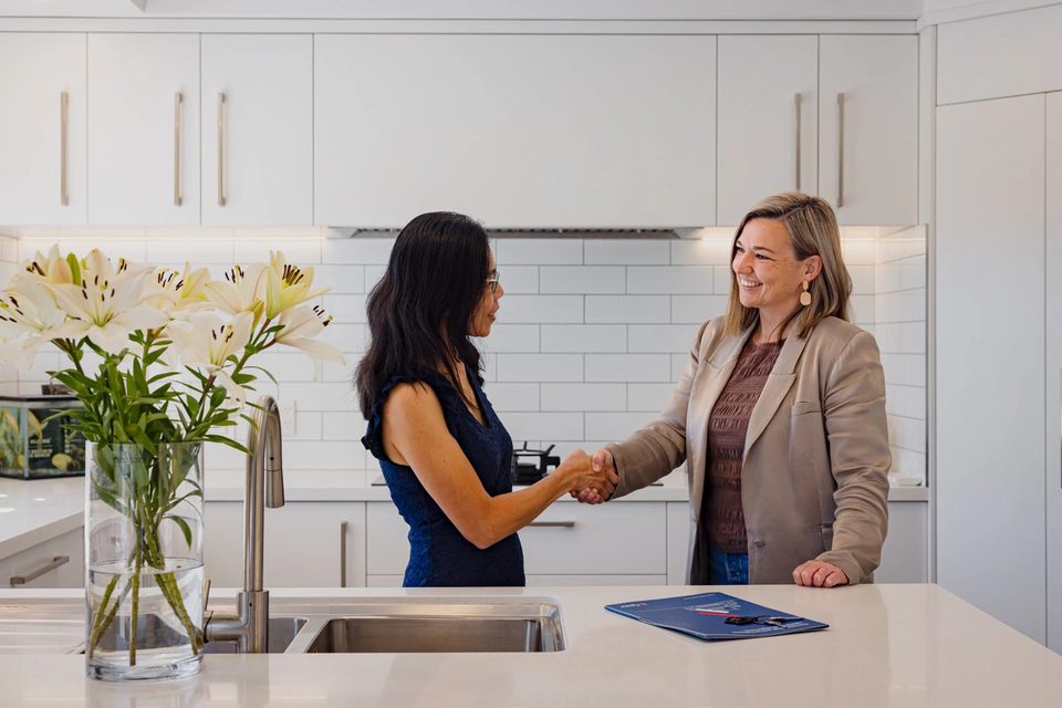 Two women shaking hands across a kitchen bench, a blue Pukeko management folder beside them.