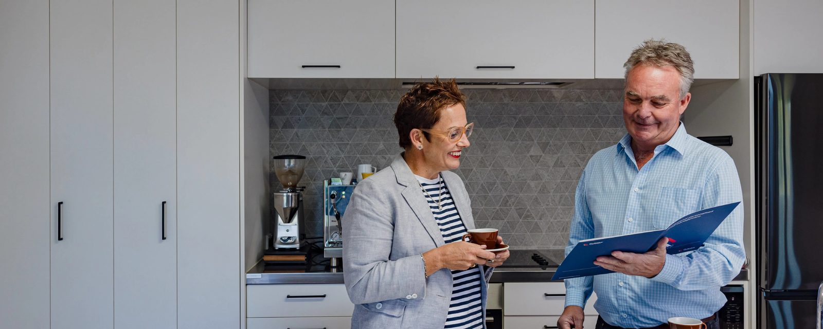 A Pukeko rental manager with a coffee cup talking to a property owner holding a blue Pukeko folder in a kitchen.