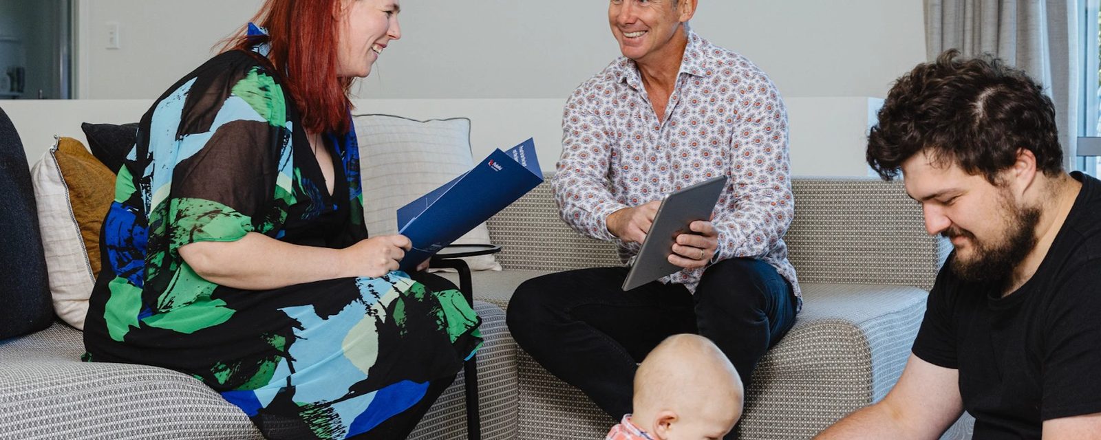 A Pukeko rental manager holding a blue folder talking with a couple and their baby in a lounge.