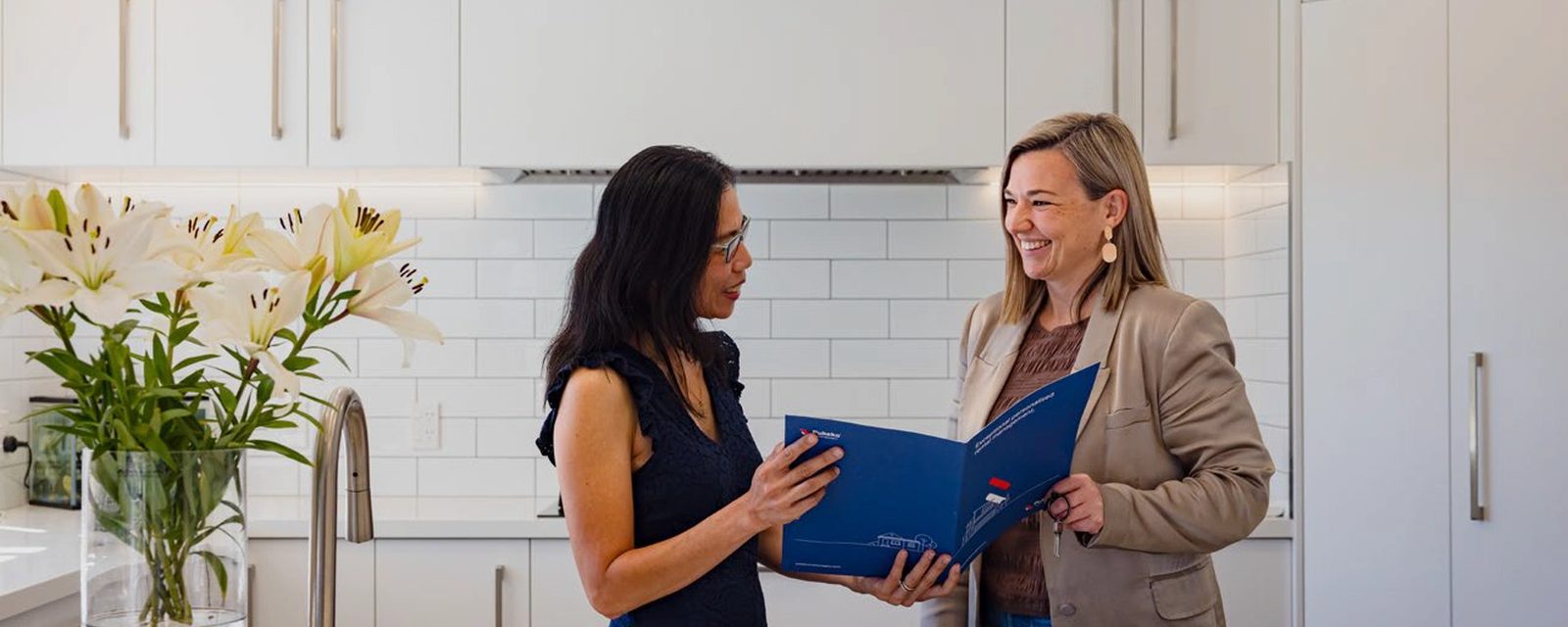 Two women reading a blue Pukeko management folder together in a white kitchen.