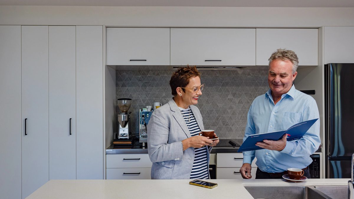 A Pukeko rental manager with a coffee cup talking to a property owner holding a blue management folder in a kitchen.